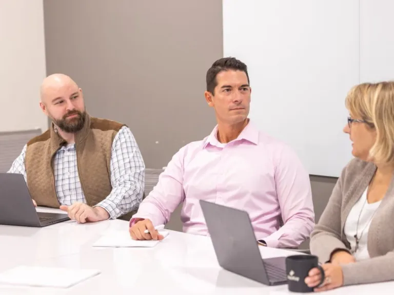 Colleagues in a meeting sit around a table with laptops, while one man takes notes