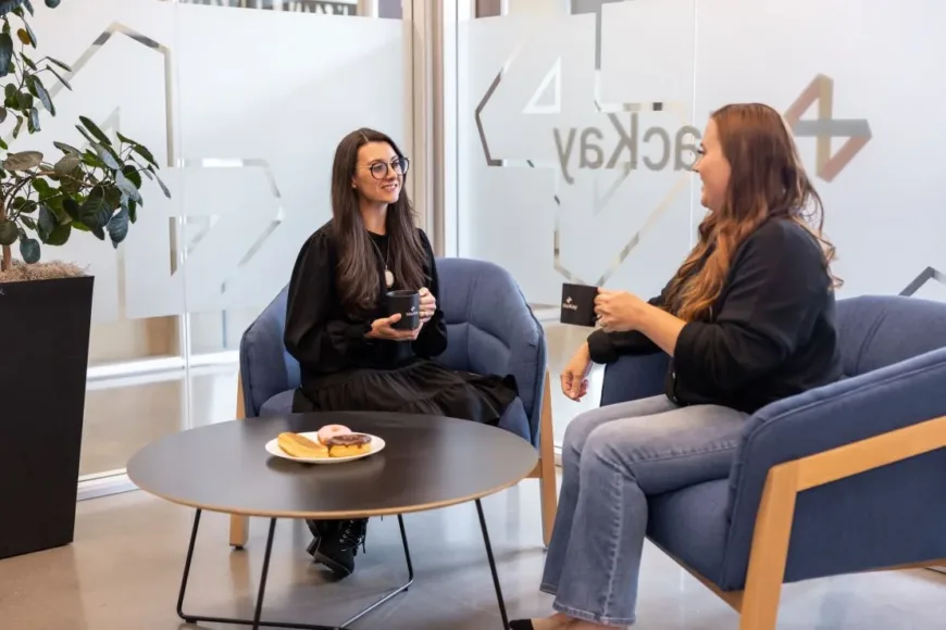 Two women chat over coffee in a modern lounge, seated in blue armchairs beside a table with pastries