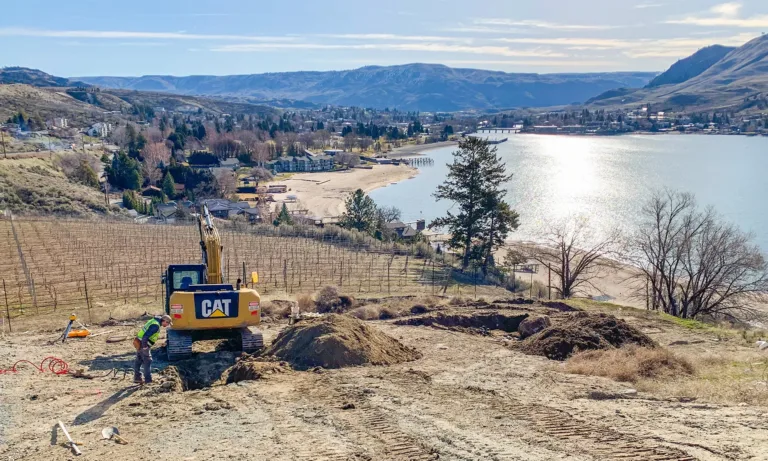 Worker beside a CAT excavator on a vineyard hillside overlooking a lakeside town and surrounding mountains