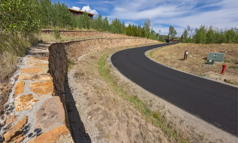 Newly paved road curves past terraced stone retaining walls and wooded hills under a blue sky