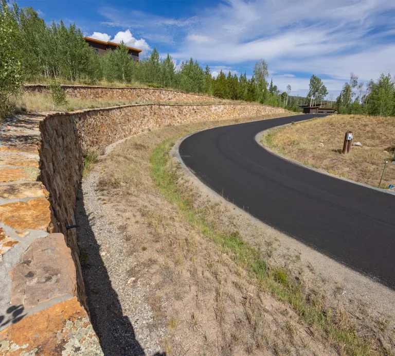 Newly paved road curves past terraced stone retaining walls and wooded hills under a blue sky