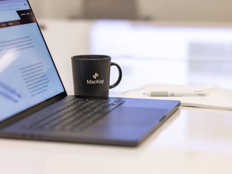 Black MacKay-branded mug beside an open laptop, papers, and a white pen on a desk