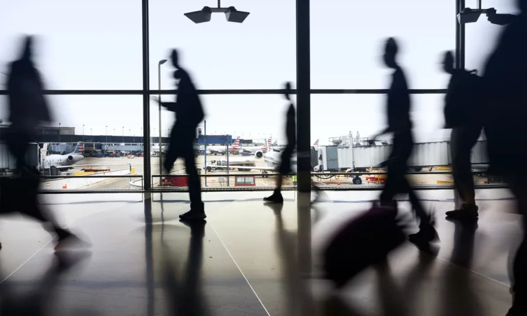 Blurred travelers walk through an airport terminal, with airplanes visible on the tarmac beyond the windows