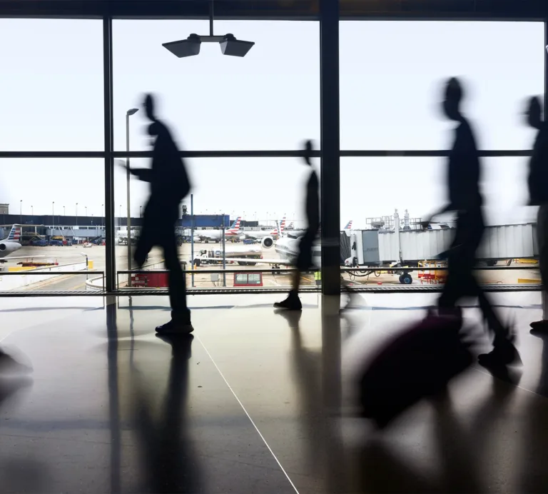 Blurred travelers walk through an airport terminal, with airplanes visible on the tarmac beyond the windows