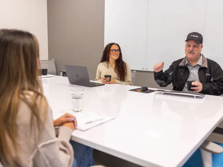 Two people sit at a conference table with laptops and coffee, appearing to discuss a presentation