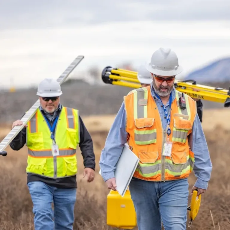 Two surveyors in hard hats and high-visibility vests carry surveying equipment through a field