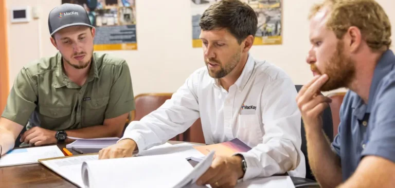 Three men review documents together around a table in a MacKay office