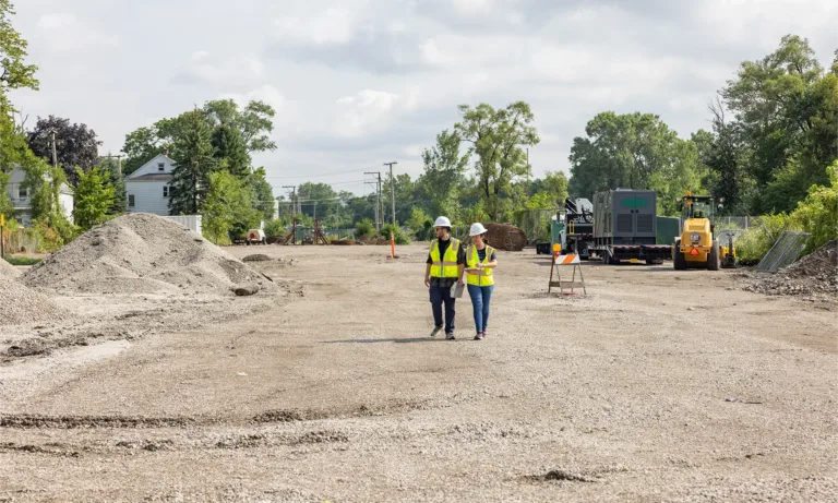 Two construction workers in high-visibility vests walk through a roadwork site with heavy equipment and gravel piles