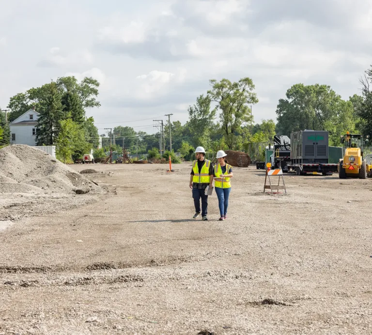 Two construction workers in high-visibility vests walk through a roadwork site with heavy equipment and gravel piles