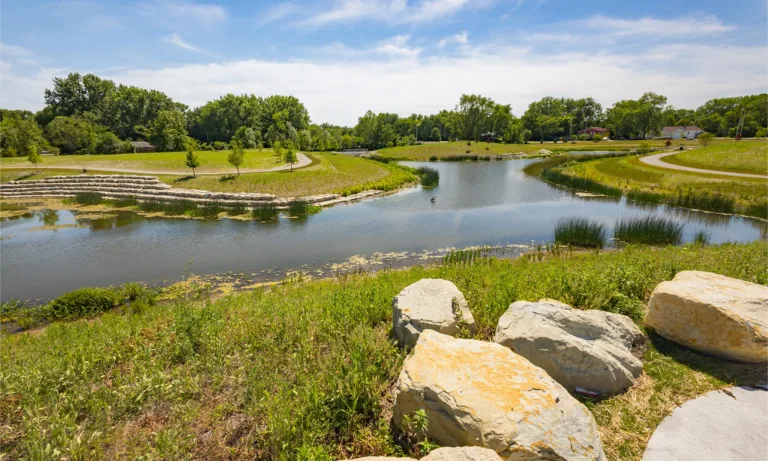 Park pond with walking paths, landscaped stone terraces, native vegetation, and large rocks along the shoreline