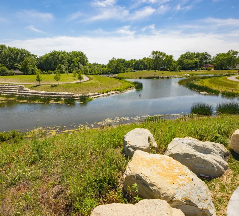 Park pond with walking paths, landscaped stone terraces, native vegetation, and large rocks along the shoreline