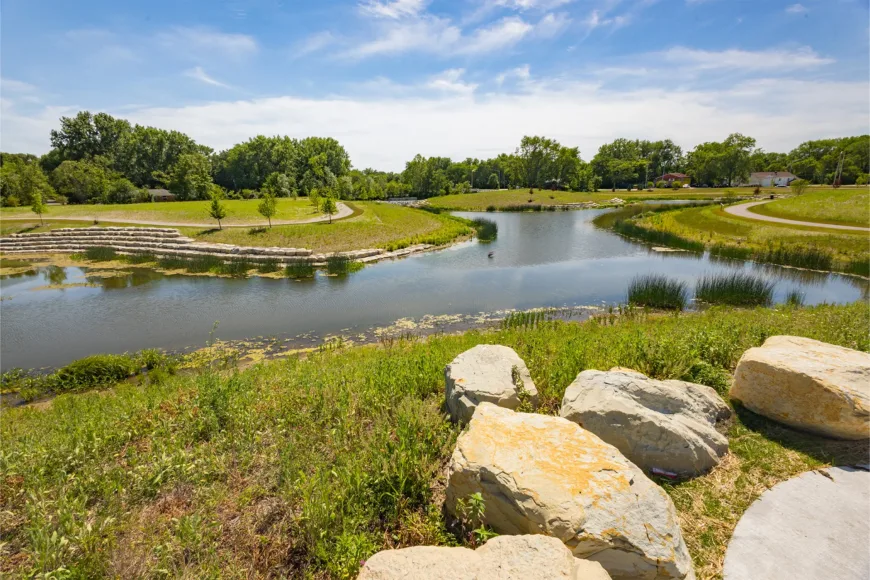 Park pond with walking paths, landscaped stone terraces, native vegetation, and large rocks along the shoreline