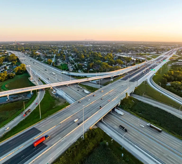 Aerial view of a busy multi-level highway interchange at sunset, surrounded by trees and residential neighborhoods
