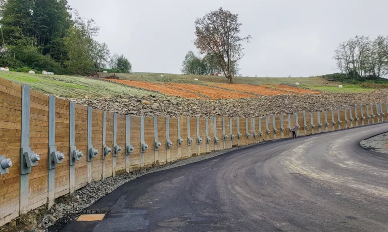 Newly paved road beside a reinforced retaining wall, with a grassy, tree-lined embankment and white hay bales beyond