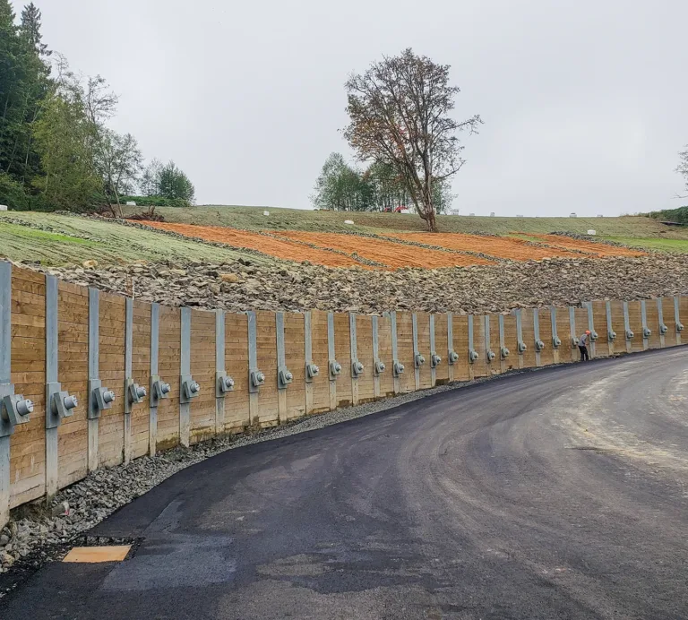 Newly paved road beside a reinforced retaining wall, with a grassy, tree-lined embankment and white hay bales beyond