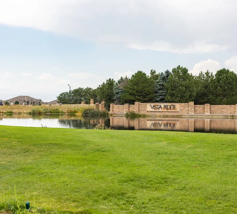 Vista Ridge community entrance beside a pond, with a stone sign, homes, trees, and landscaped lawn