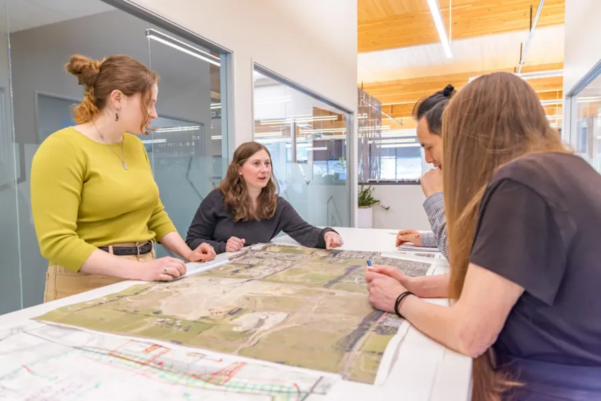 Four colleagues discuss plans over large maps and architectural drawings in an office
