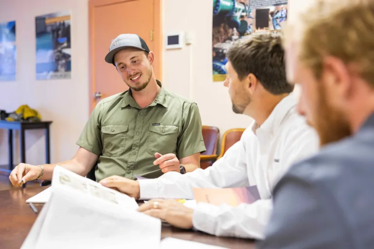 Three men sit around a table, reviewing papers and discussing them in an office