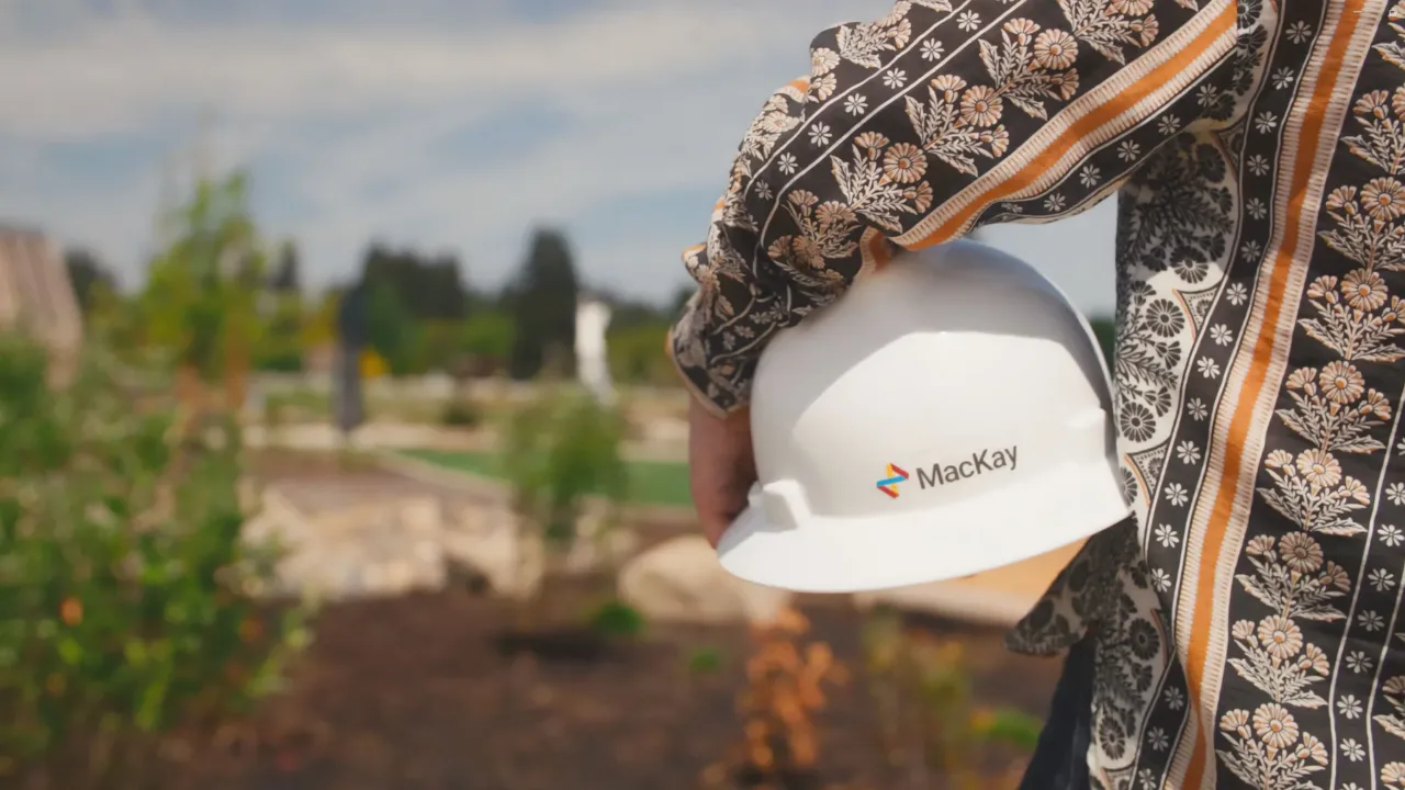 Person holding a white hard hat labeled “MacKay” at an outdoor worksite
