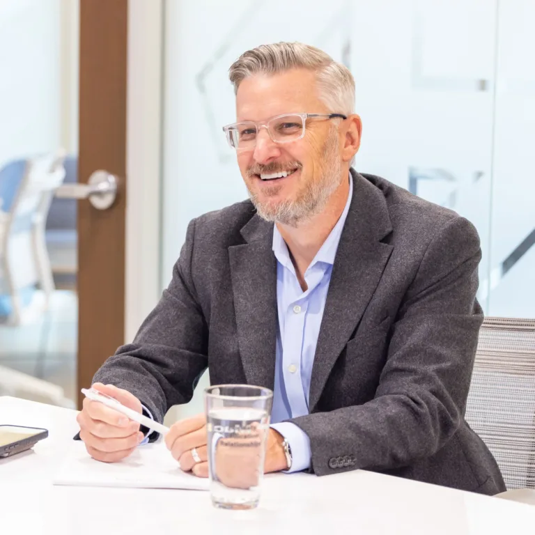 Smiling man in glasses and a blazer writing at a meeting table with a glass of water