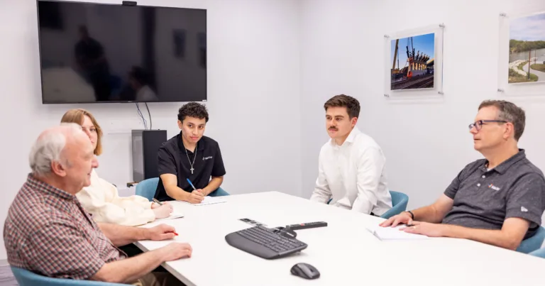 A diverse team gathers around a conference table for a meeting, with one participant taking notes