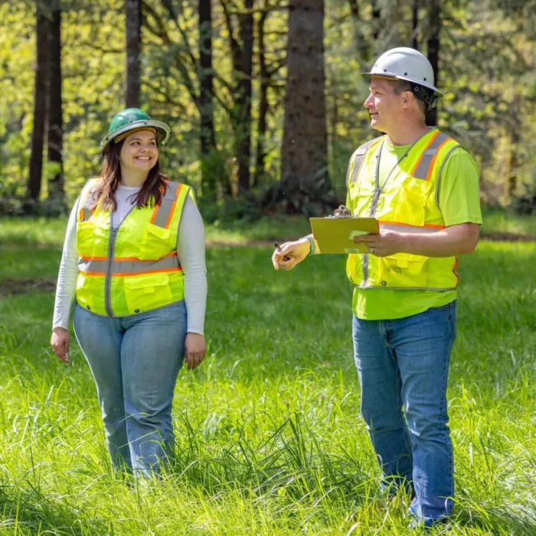 Two forestry workers in high-visibility vests and hard hats stand in a grassy forest, one holding a clipboard