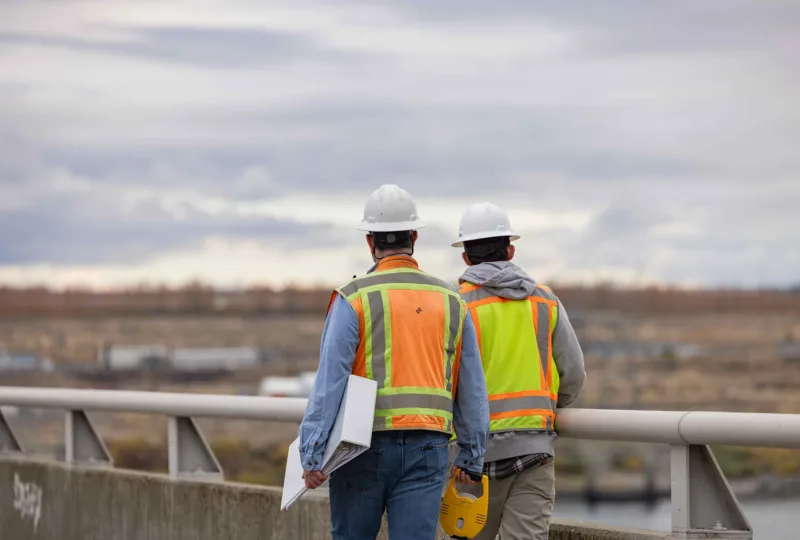 Two construction workers in hard hats and high-visibility vests stand on a bridge overlooking an industrial site