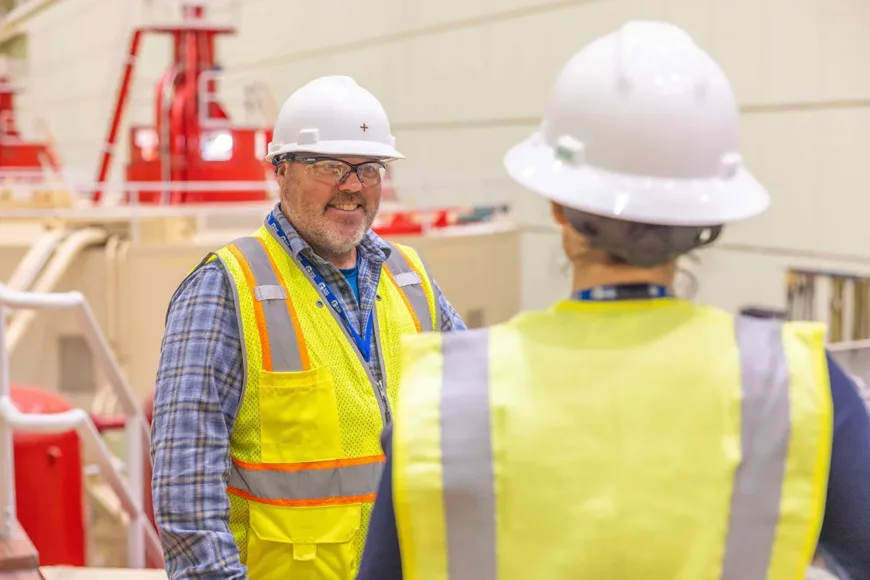 Two construction workers in hard hats and high-visibility vests converse inside an industrial facility