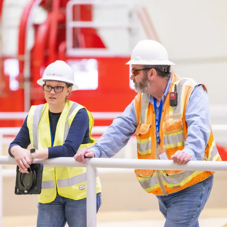 Two industrial workers in hard hats and high-visibility vests inspect a facility from behind a safety railing
